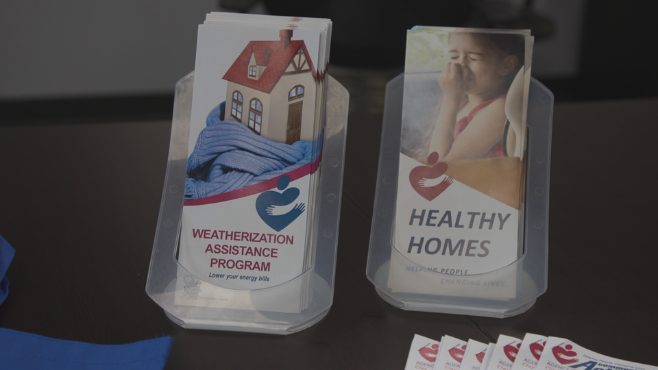 Two stacks of pamphlets on a table. The table is brown. The pamphlets to the left of the screen advertise a weatherization assistance program. These pamphlets have a house with a red roof on them wrapped in a blue scarf. The text is bold and red. The pamphlets on the right advertise the healthy homes program. On these pamphlets, a young girl holds her nose. The text on these pamphlets is black and bold.