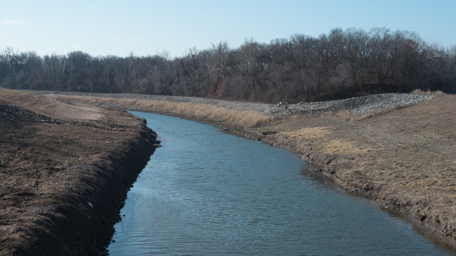 A photo of the Blue River. The river is larger at the foreground of the image and thins to a point in the background of the image. The water is blue with small ripples. On the sides of the river are clean, grassy banks. Just beyond the river bend in the background of the photo is a treeline.