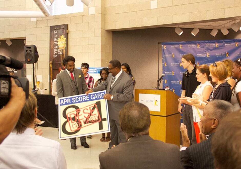 Camera and audience face podium where students watch as KCPS Superintendent Dr. Steve Green receives a large card with the district's 84 point APR score.