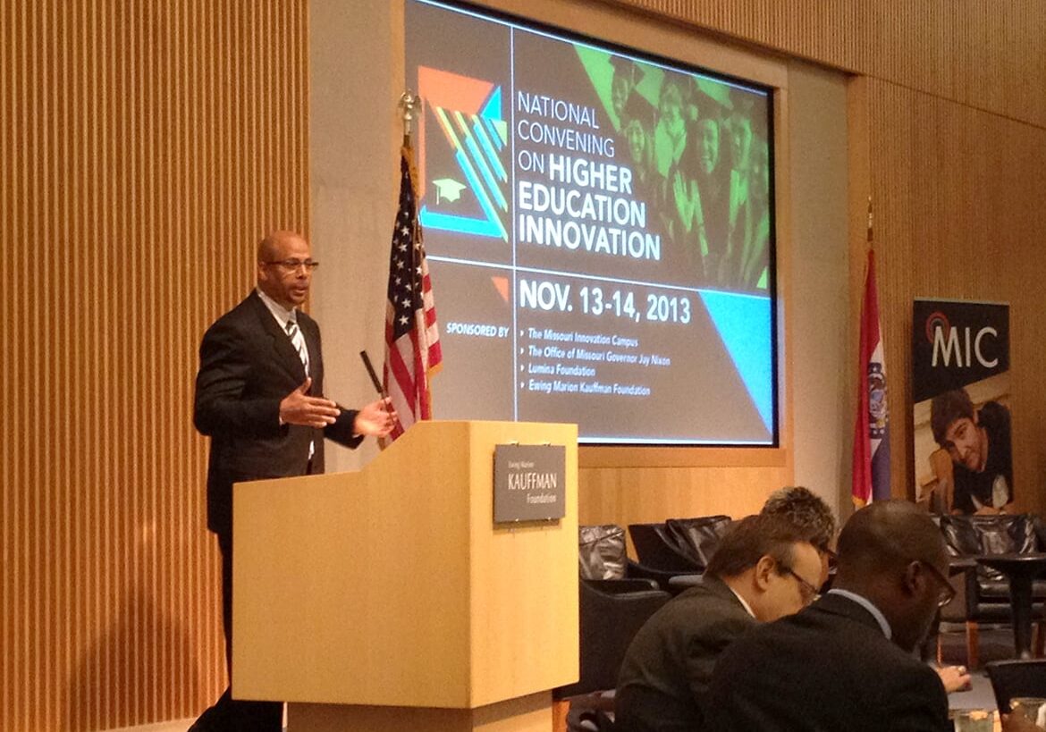 Man in suit and tie standing at podium in front of screen that reads "National Convening on Higher Education Innovation"
