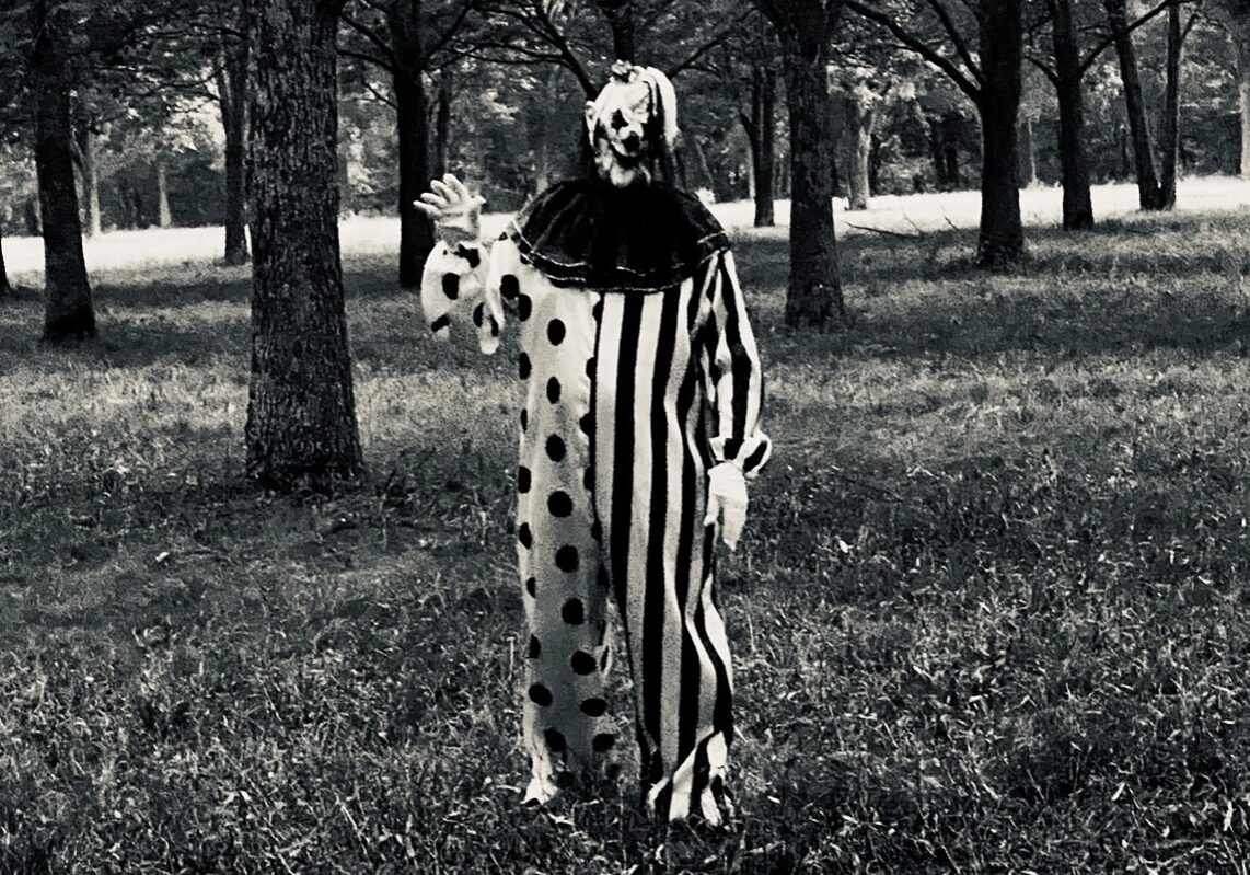 A clown stands in a field in a creepy outfit.