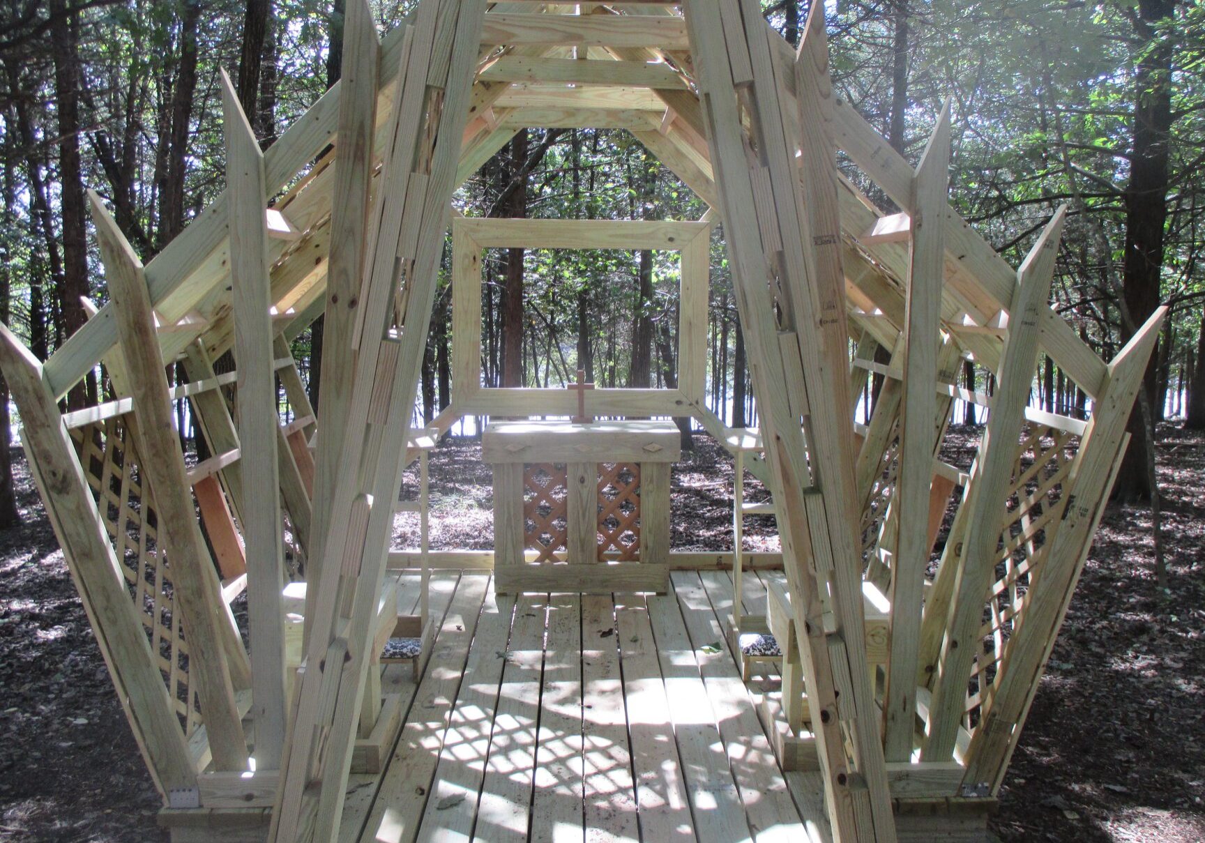 A small wooden Structure shaped like a chapel sits in the woods with dappled sunlight on its floor. Inside is an altar with a cross.