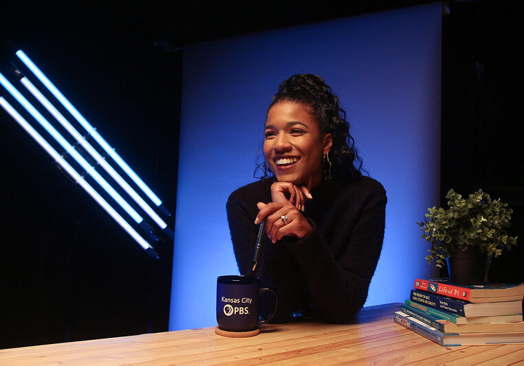 woman sits in front of a blue background. She has a coffee cup that reads "Kansas City PBS" and a stack of books with a plant next to here.
