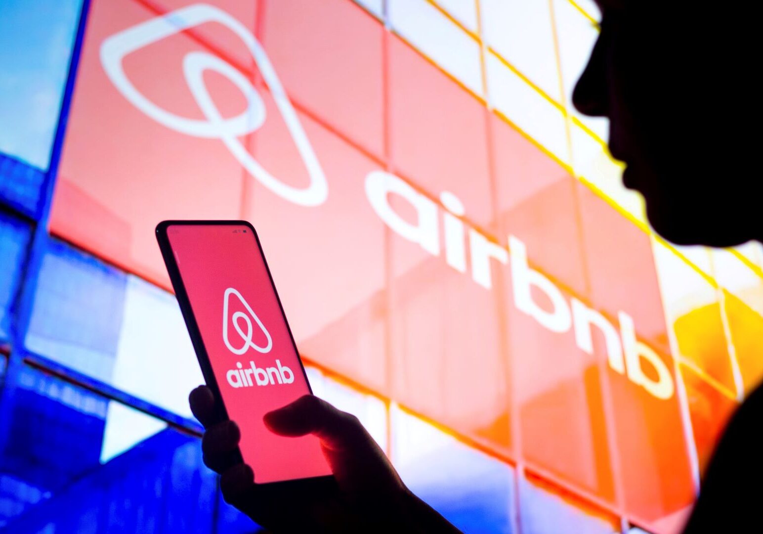 In this photo illustration, a woman's silhouette holds a smartphone with the Airbnb logo displayed on the screen and in the background.