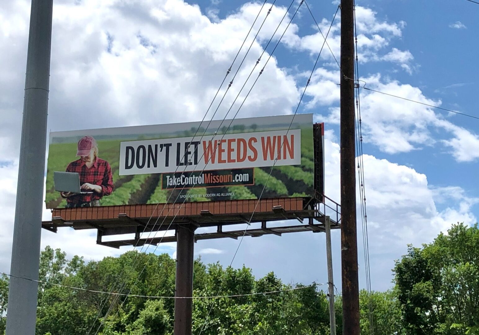 A billboard with a farmer holding a laptop in rows of a crop. Text on the billboard reads "Don't let weeds win" and "takecontrolmissouri.com"
