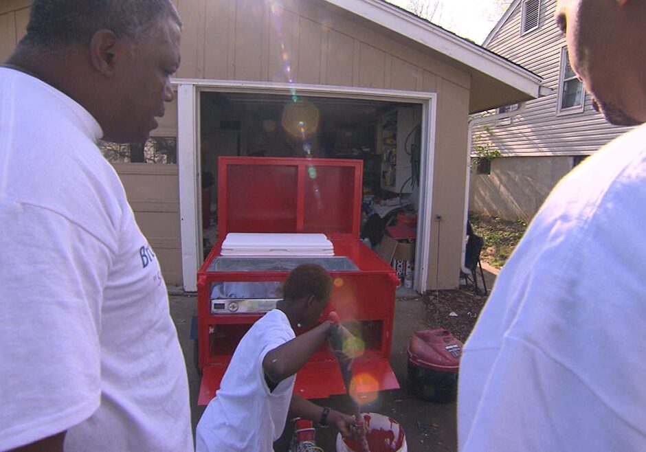 Two men watch as another paints a hot dog cart in front of a garage.