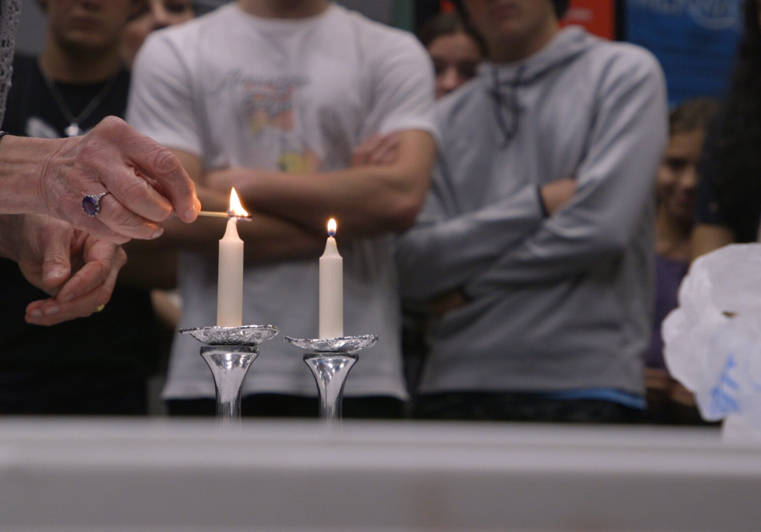 A elderly woman holds a match over a white candlestick. She's wearing a large amethyst ring. High schoolers watch in the background.