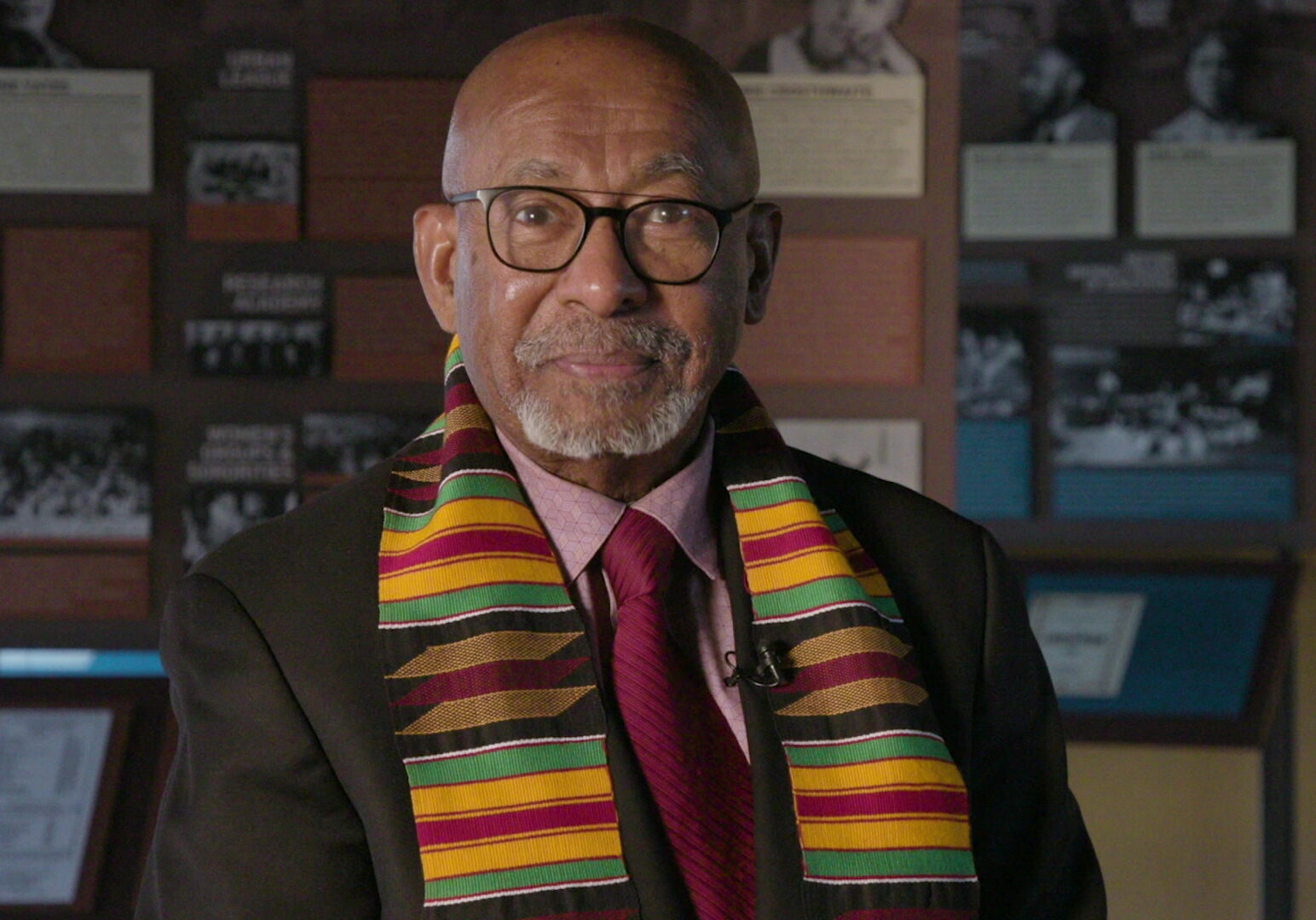 A man with white facial hair and classes sits in front of a display with historical documents. He wears a traditional scarf with reds, yellows, blacks and greens.