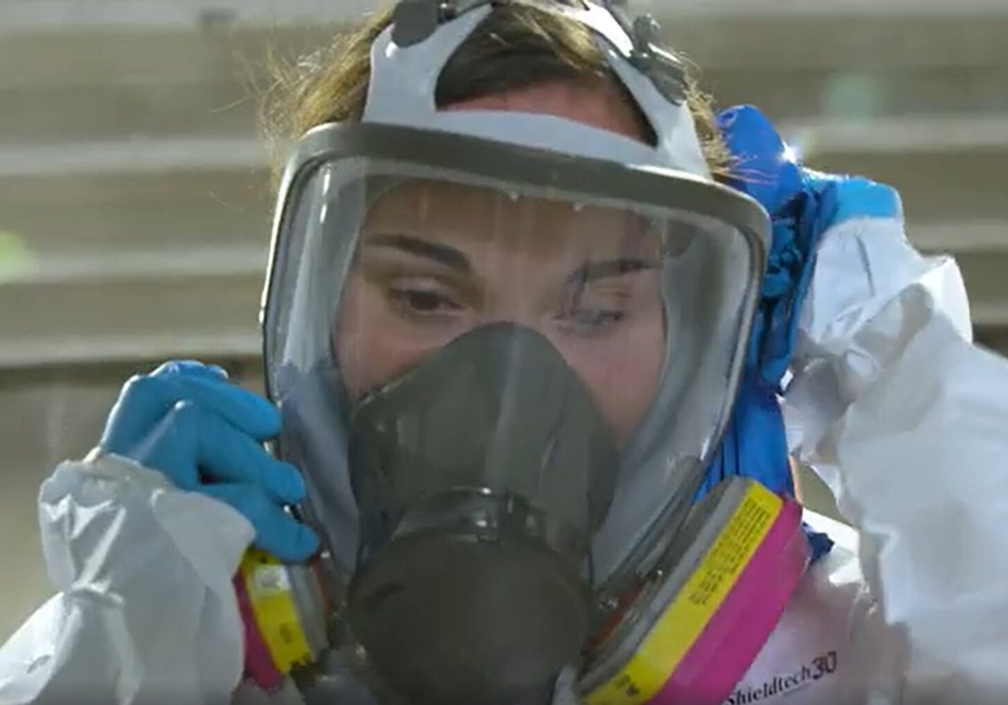 A women in a white hazmat suit puts on a mask.