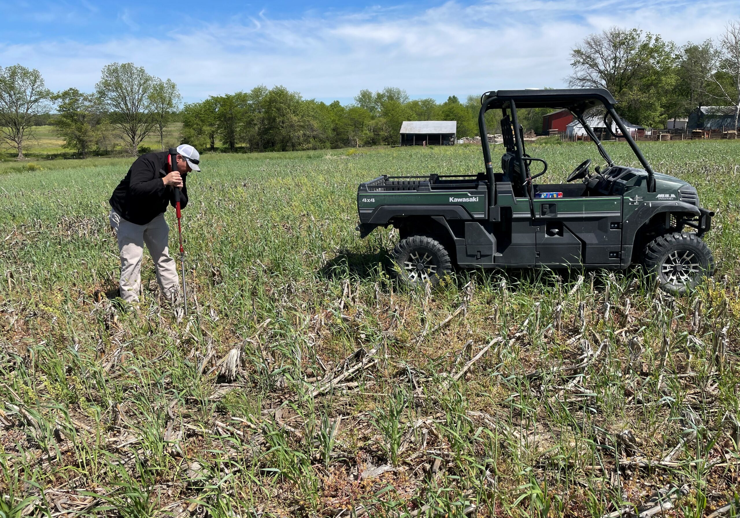 A man stands in a field that has dead corn stalks and a young cover crop. He is digging for a soil sample. Next to him is a green four wheeler.