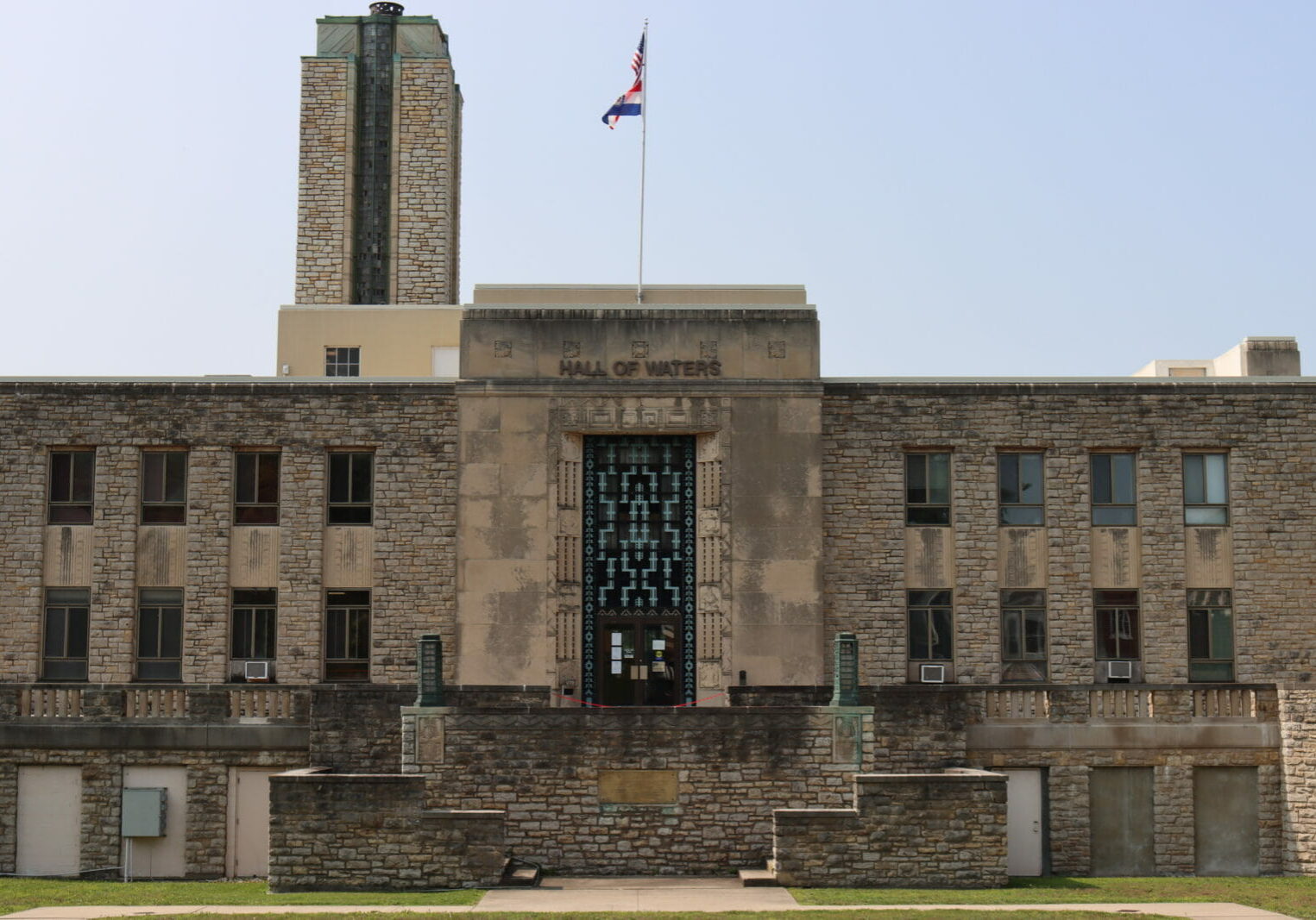 A stone building with bluish green metal on the front reads "Hall of Waters" a stone water tower is on top of the building, as is a flag.