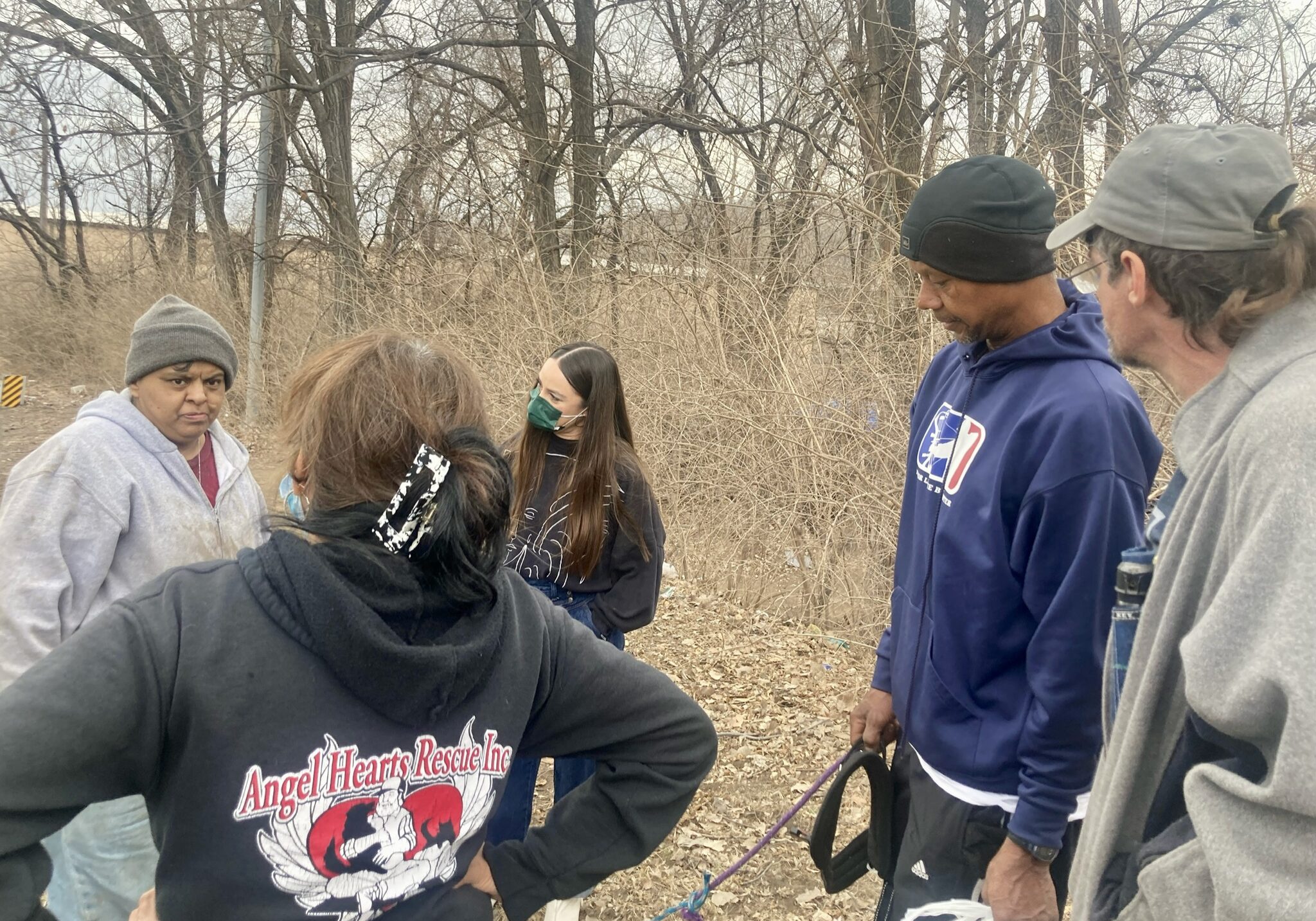 A group of people stand outside. Several people belong to an encampment where people experiencing homelessness live. One woman Alice Delbosque has her back to the camera. She is wearing a sweatshirt of her organization called Angel Hearts Rescue, which provides aid to pets belonging to those experiencing homelessness.
