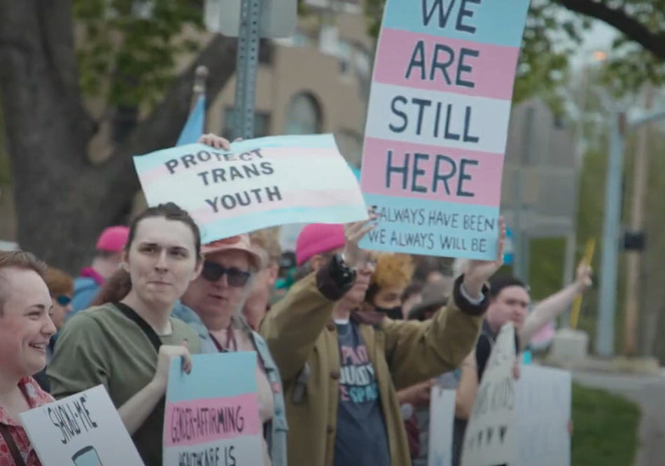 Folks hold up pink white and blue signs that read "We Are Still Here" at a transgender rights protest in Kansas City.
