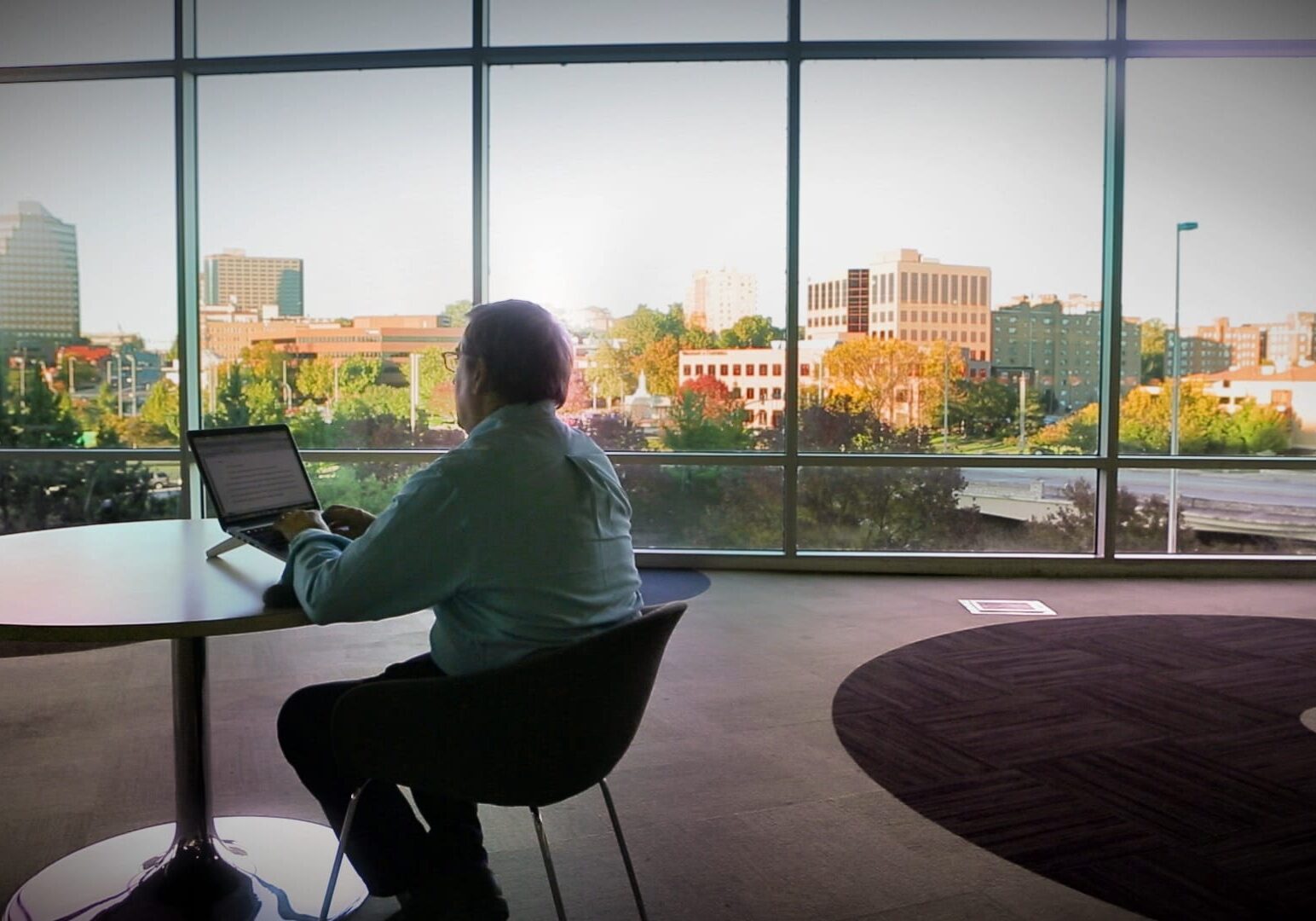 Image of man working on computer with mid-town Kansas City buildings outside large windows