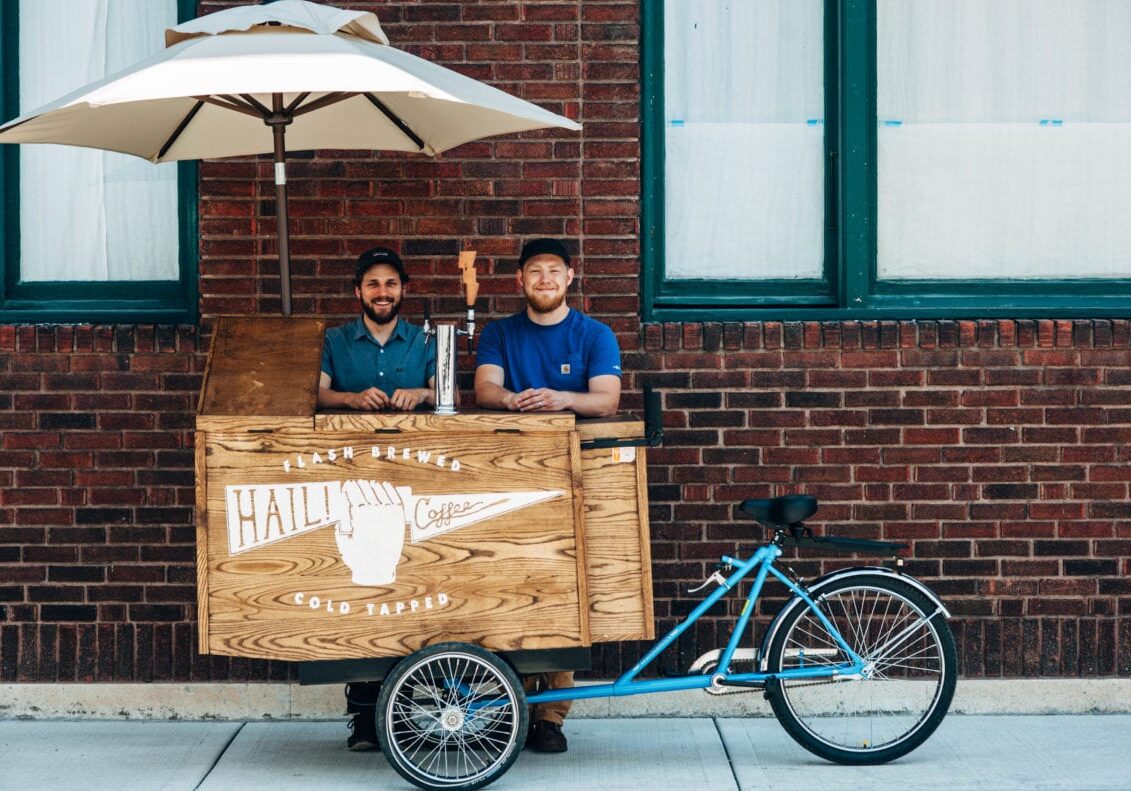Two men stand behind their coffee cart.
