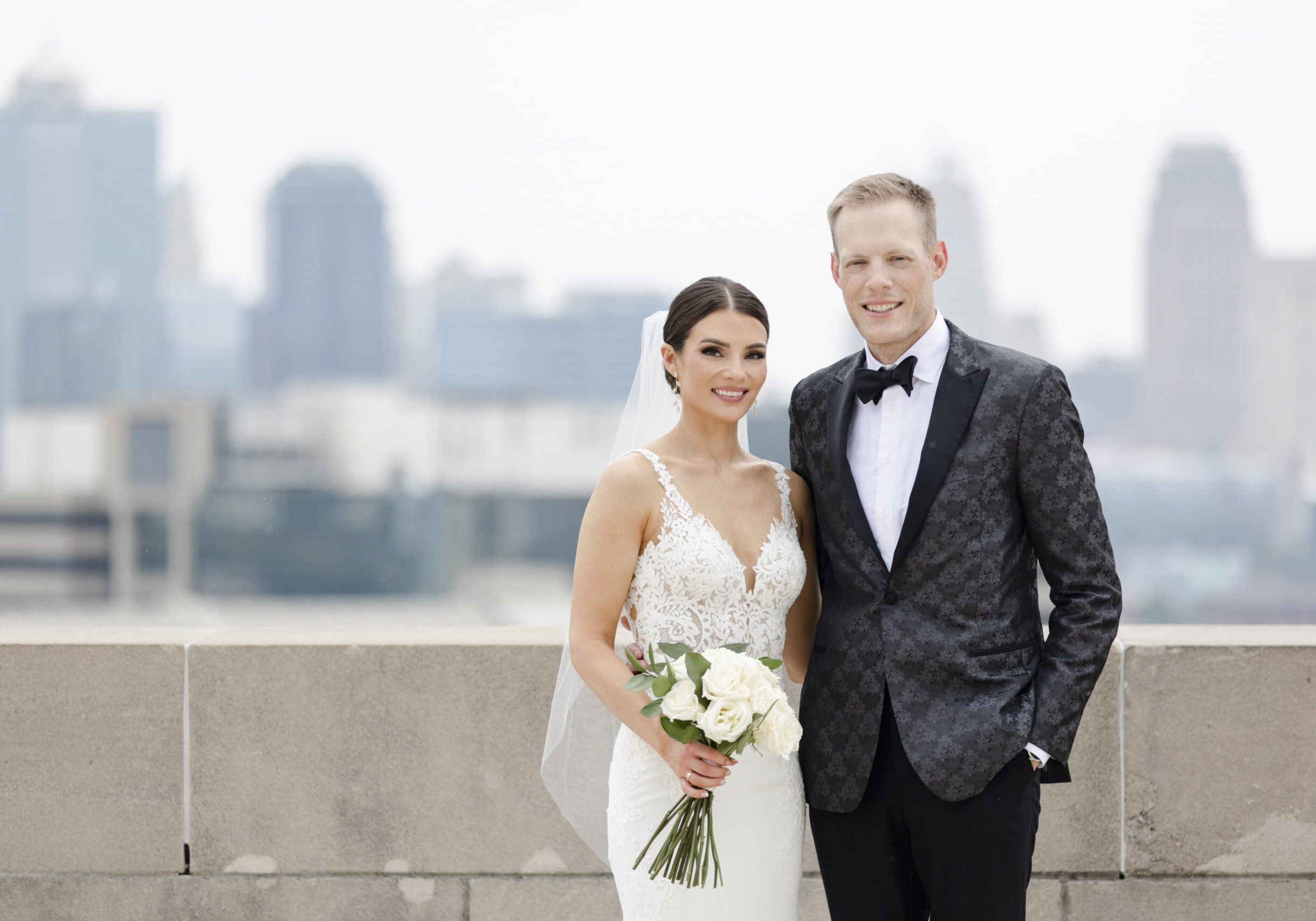 A wedding portrait. A young white couple, woman wearing a white gown holding a bouquet, and man in a tuxedo, stand outside of Kansas City's citscape. Joey and Audrey Sughroue got married July 2022. Their hope this year is to find a living donor for Joey, whose kidneys are at 10% function. (Contributed image)