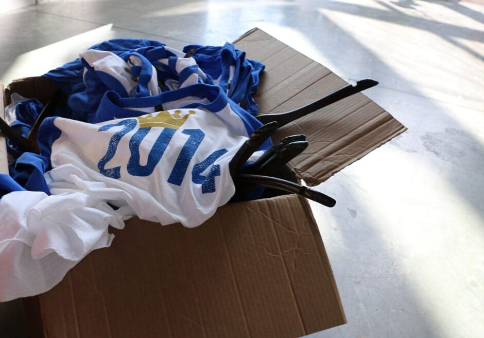 a pile of blue and white baseball T-shirts with "2014" (with a crown over the 0) overflow out of a box on a concrete floor.