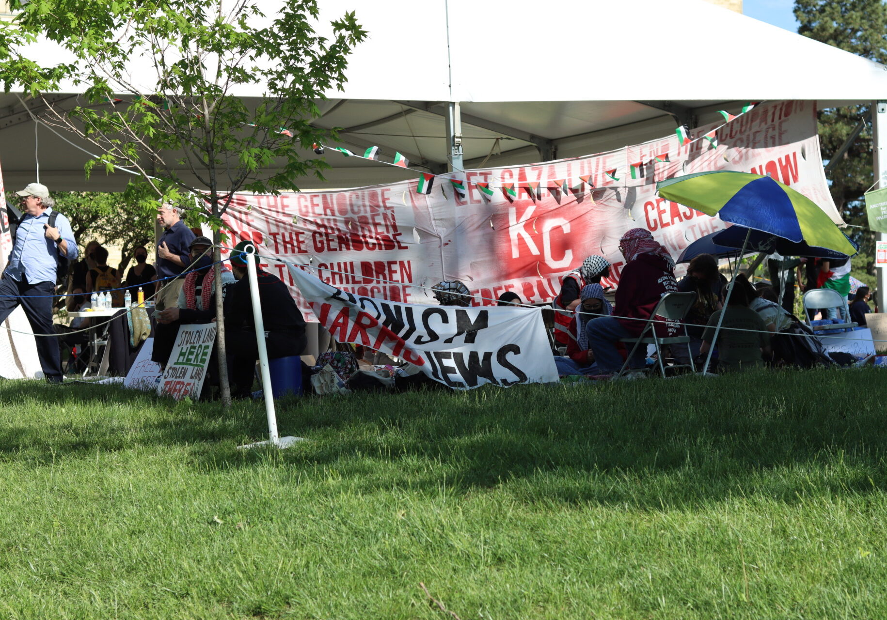 A large white tent is covered with banners reading things like "stop the genocide" "Ceasefire now" "Let Gaza Live". Around the tent sit students wearing keffiyehs.