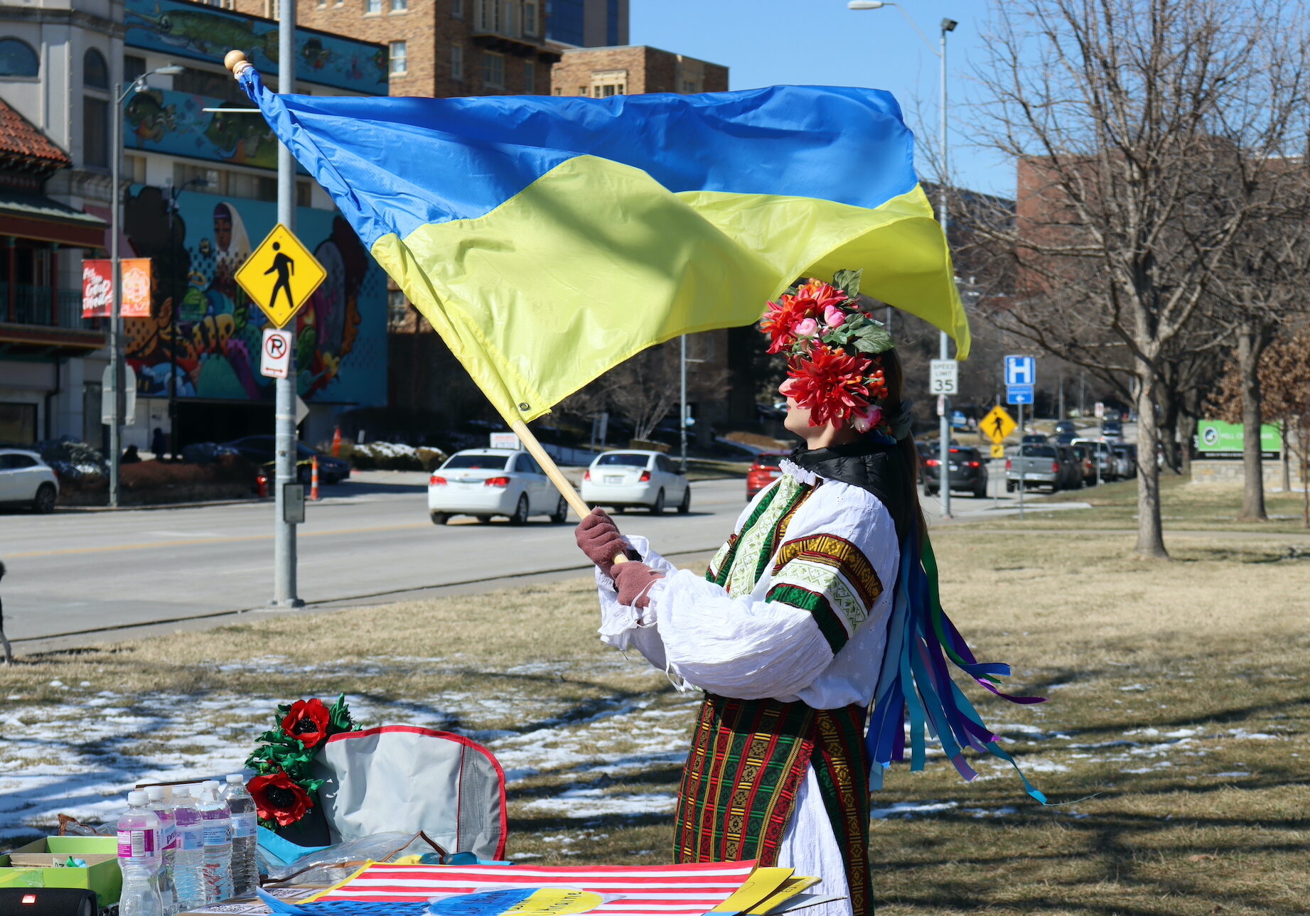 Woman in flower crown and traditional, colorful Ukrainian dress holds a blowing Ukrainian flag in a park.