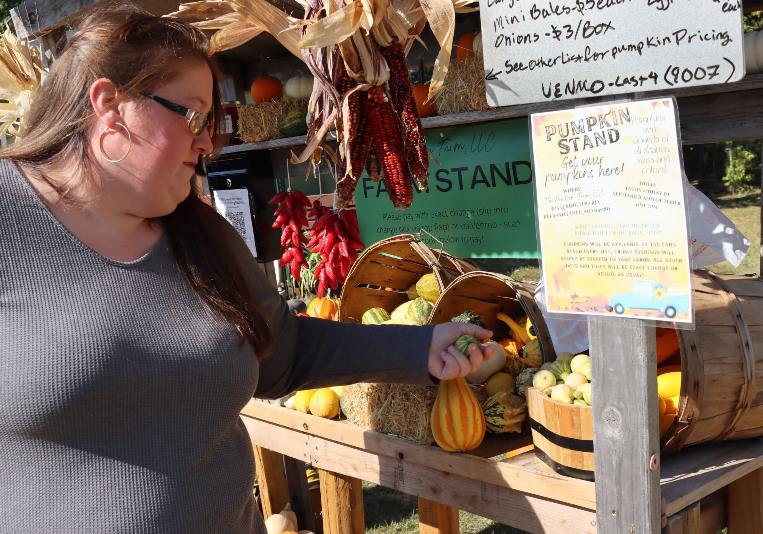 woman in gray shirt and glasses points to squash in a wooden farm stand.
