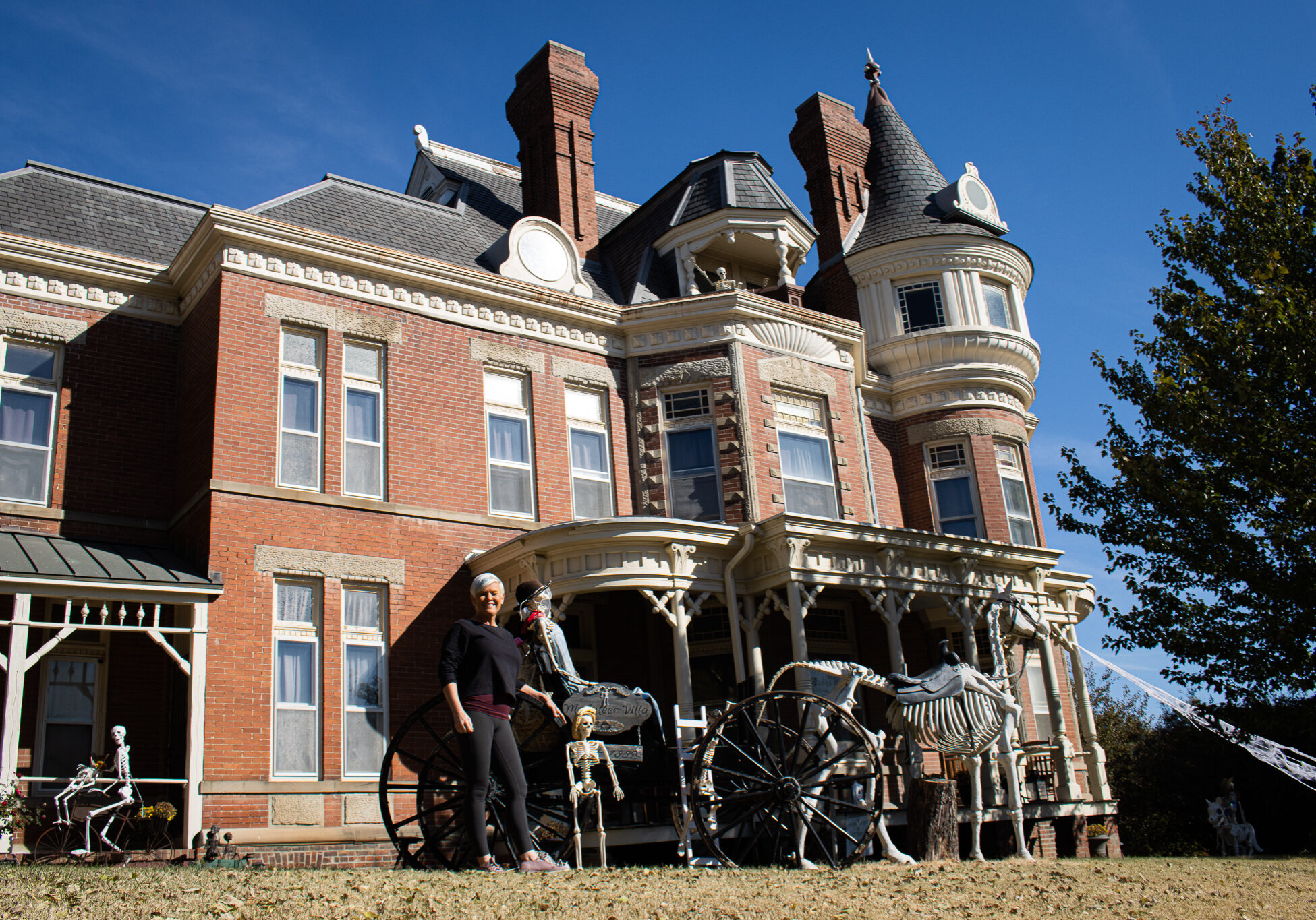 Woman stands next to old carriage with skeleton horses. Behind her is a victorian style mansion with brick facade and white trimming.