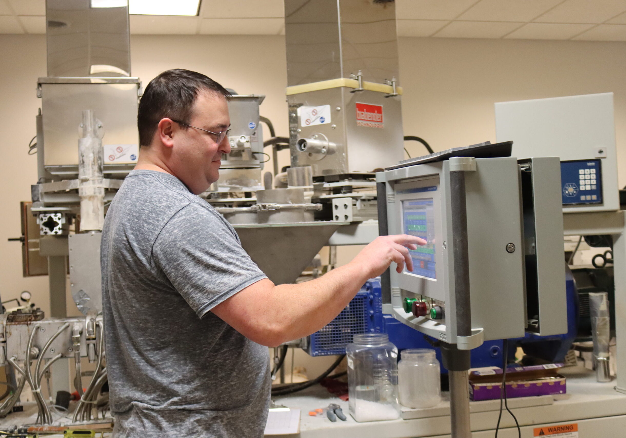 Man in a grey shirt and glasses uses the touchscreen on a large machine used for extruding plastic pellets.