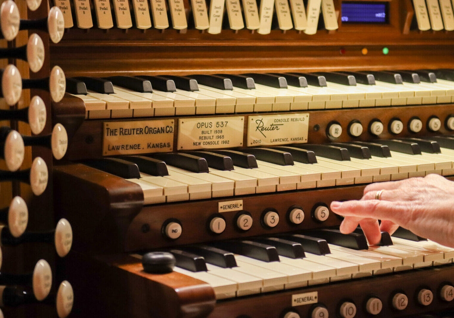 Between rows of piano keys (stacked vertically) we see plaques that read "Reuter Organ Company" A hand reaches to play several keys.