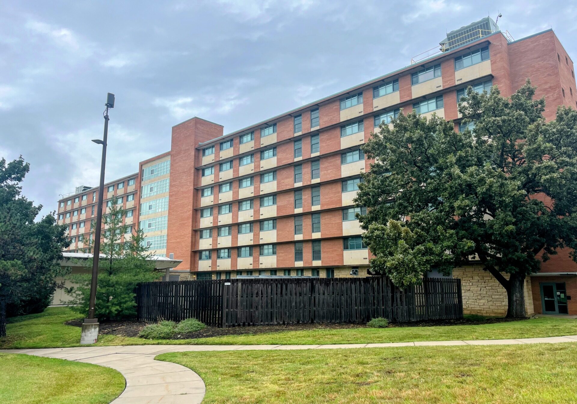 A photo of a dormitory hall. At the foreground of the photo is a green lawn with a curved sidewalk path cutting through it on the left side of the photo. The dormitory is a red color and stretches horizontally across the photo. A large tree partially covers the right side of the building.