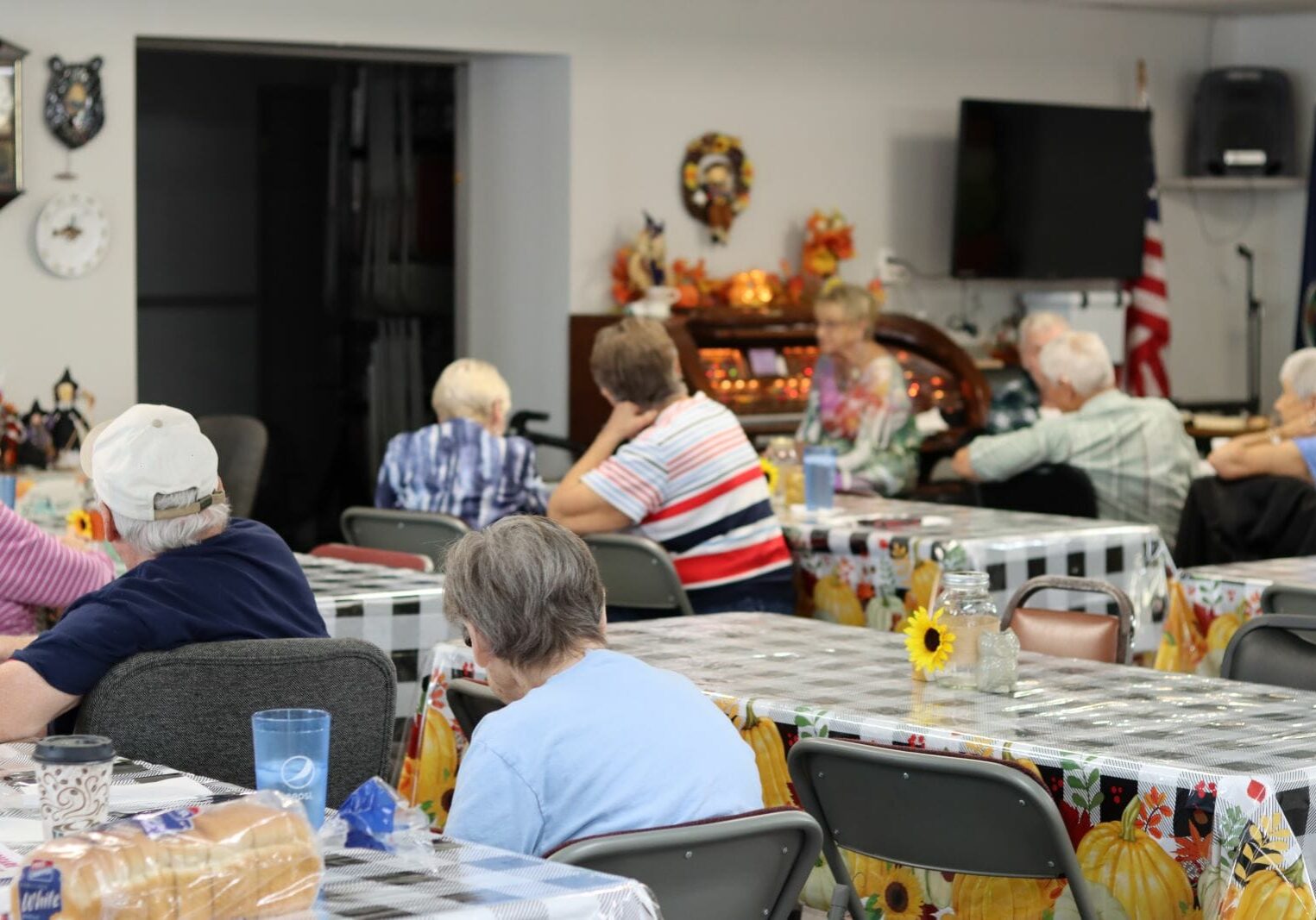 A group of older people sit at rectangular tables with checkered table cloths. In the background a woman sits at an electric organ with lots of colorful lights.