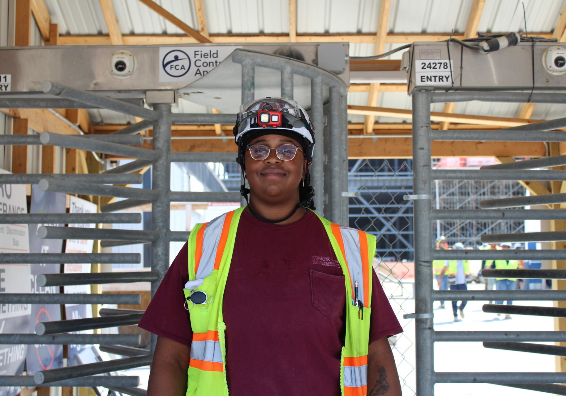 A woman wearing a dark red shirt stands in front of gray metal bars. The woman is wearing glasses, a bright yellow utility vest and a white hard hat.