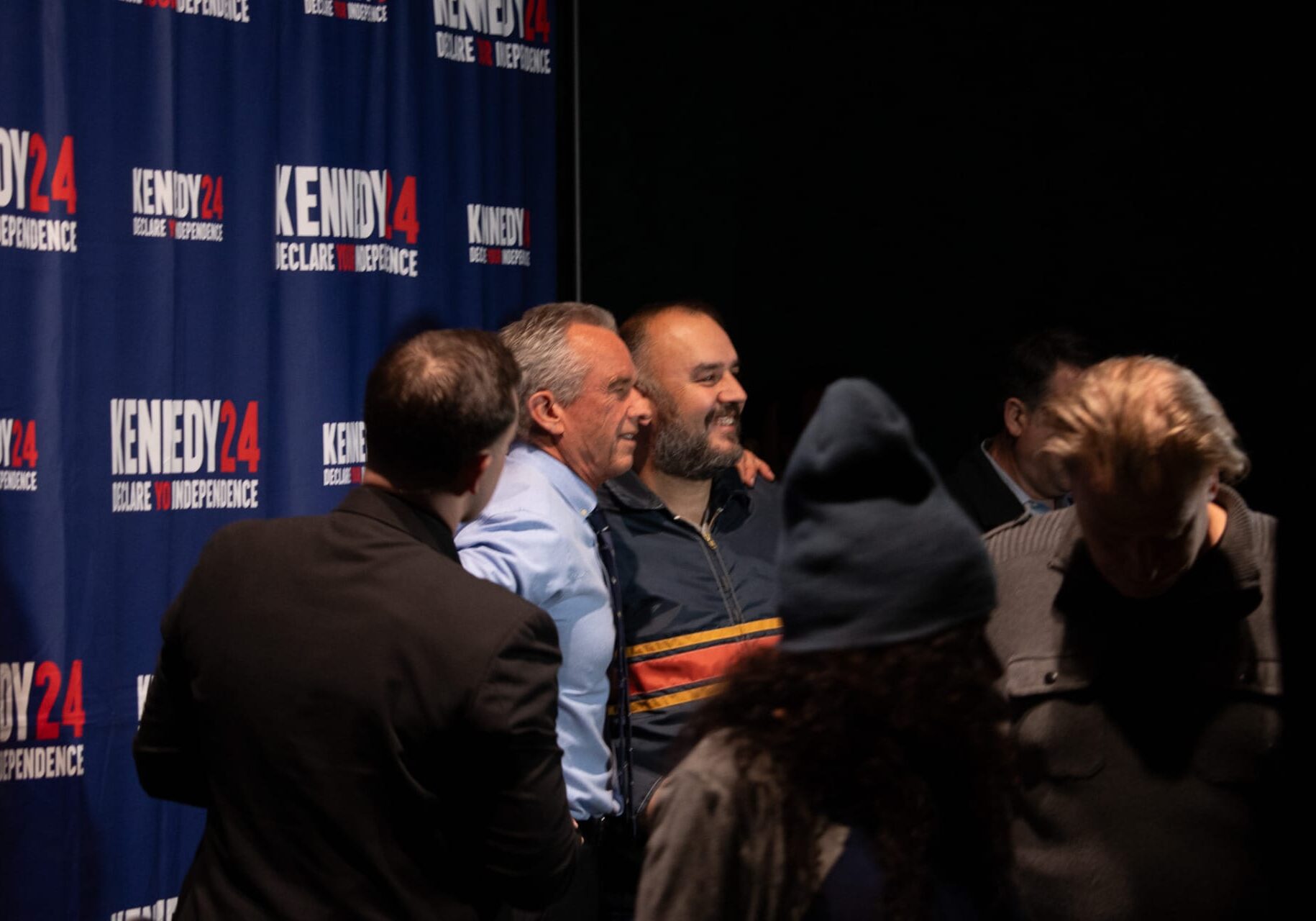 An older man in a blue shirt puts his arm around a middle aged man as they pose for a photo. They stand in front of a banner that reads "Kennedy 24 Declare Your Independence"