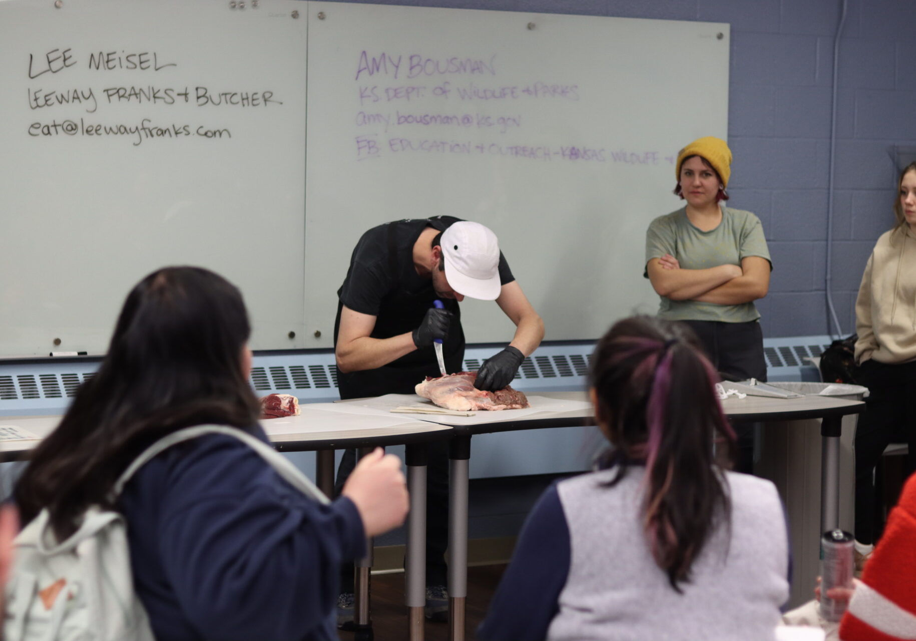 Man holding knife bends over a table cuts into slab of raw meat. People in a classroom crowd around to watch.