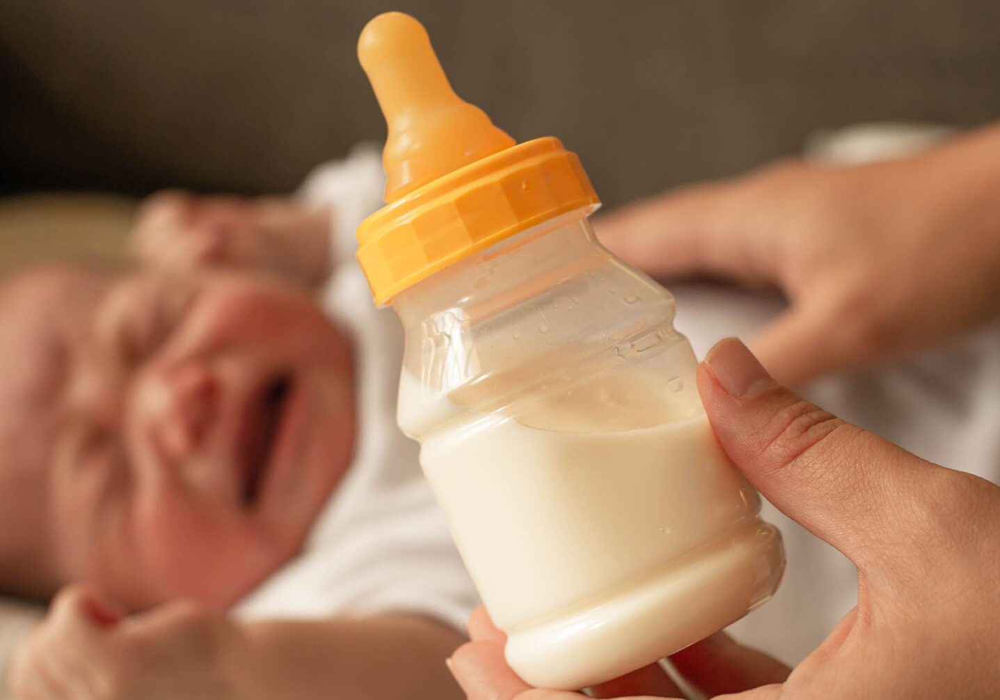 A baby bottle is held up as a baby cries in the background, illustrating the difficulty parents are having during the formula shortage. Image by Adobe Spark.