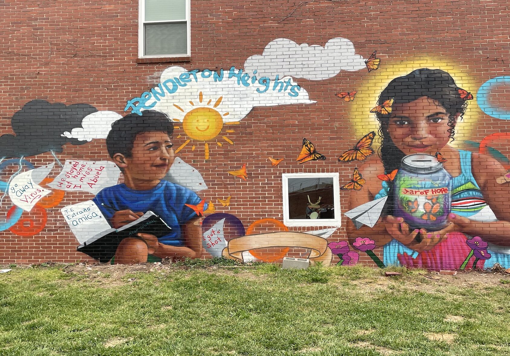 A mural of two young, Brown children painted on a red brick wall illustrates the difficulty kids felt being isolated from friends and family during the pandemic. One of the images shows a boy holding a notebook, with pages flitting around him that say "Te extraño amiga" and "go away virus."