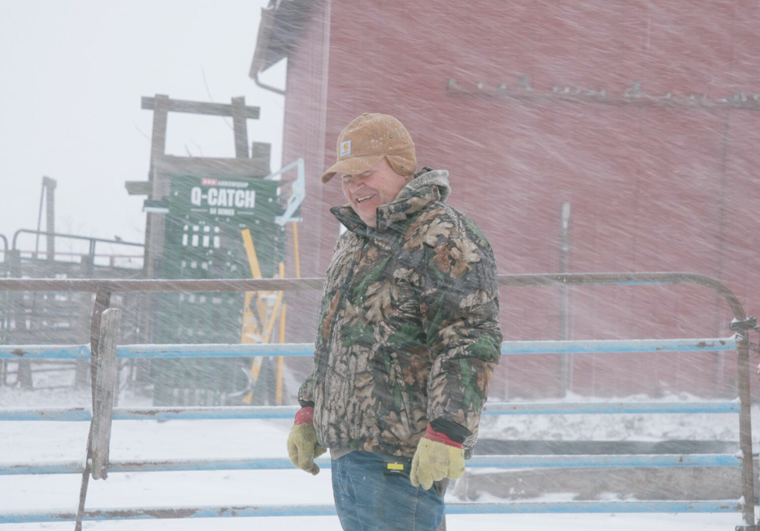 A man in a camouflage coat and a brown hat stands in front of a red barn in a blizzard.