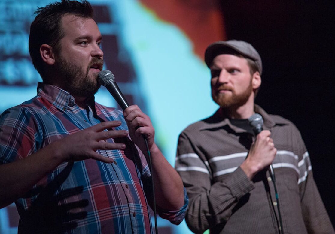 Picture of two men with microphones standing in front of a screen.