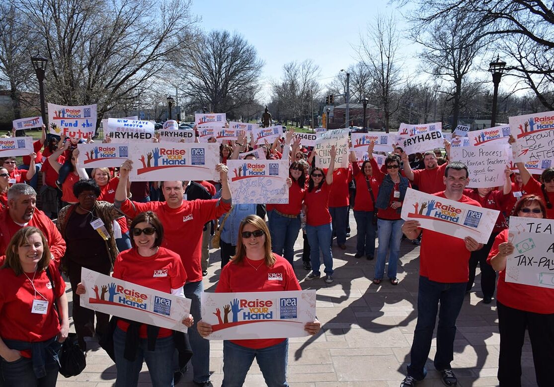 Photo of group of people outside in red shirts holding signs.