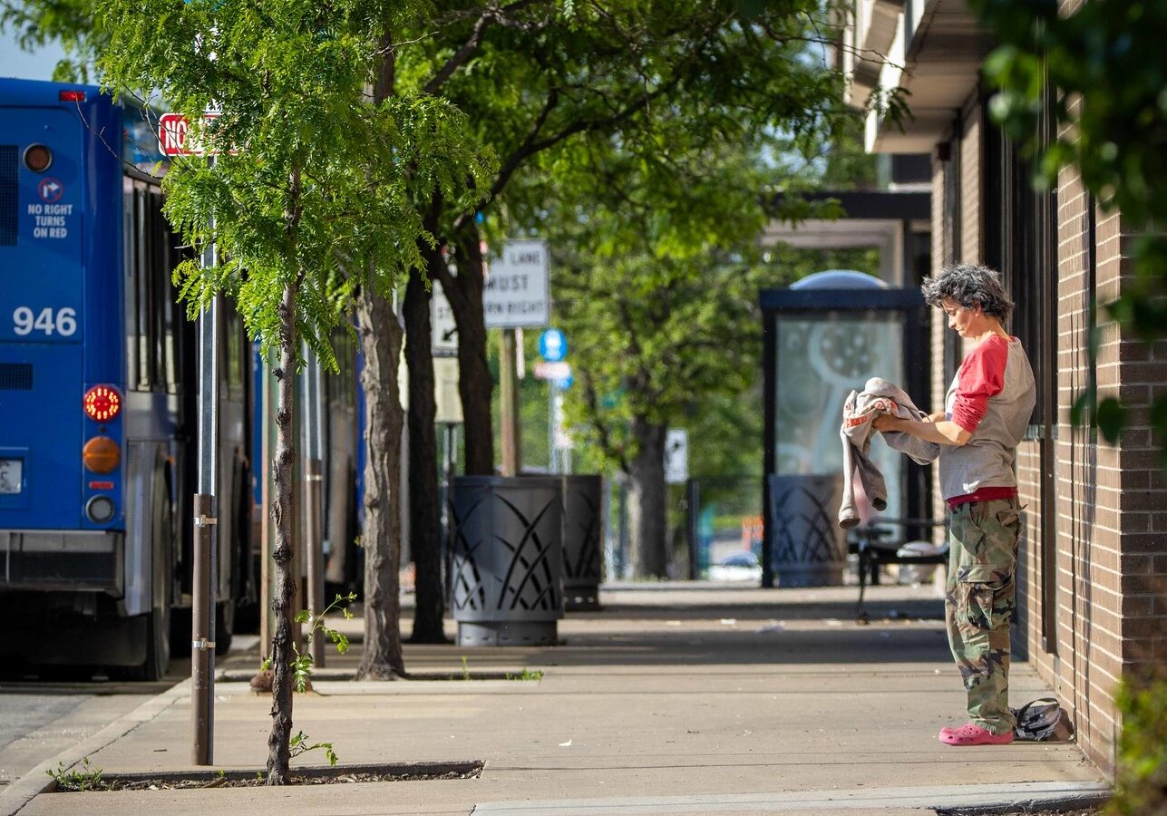 A person stands on the sidewalk near a bus stop just south of the Lawrence Public Library.