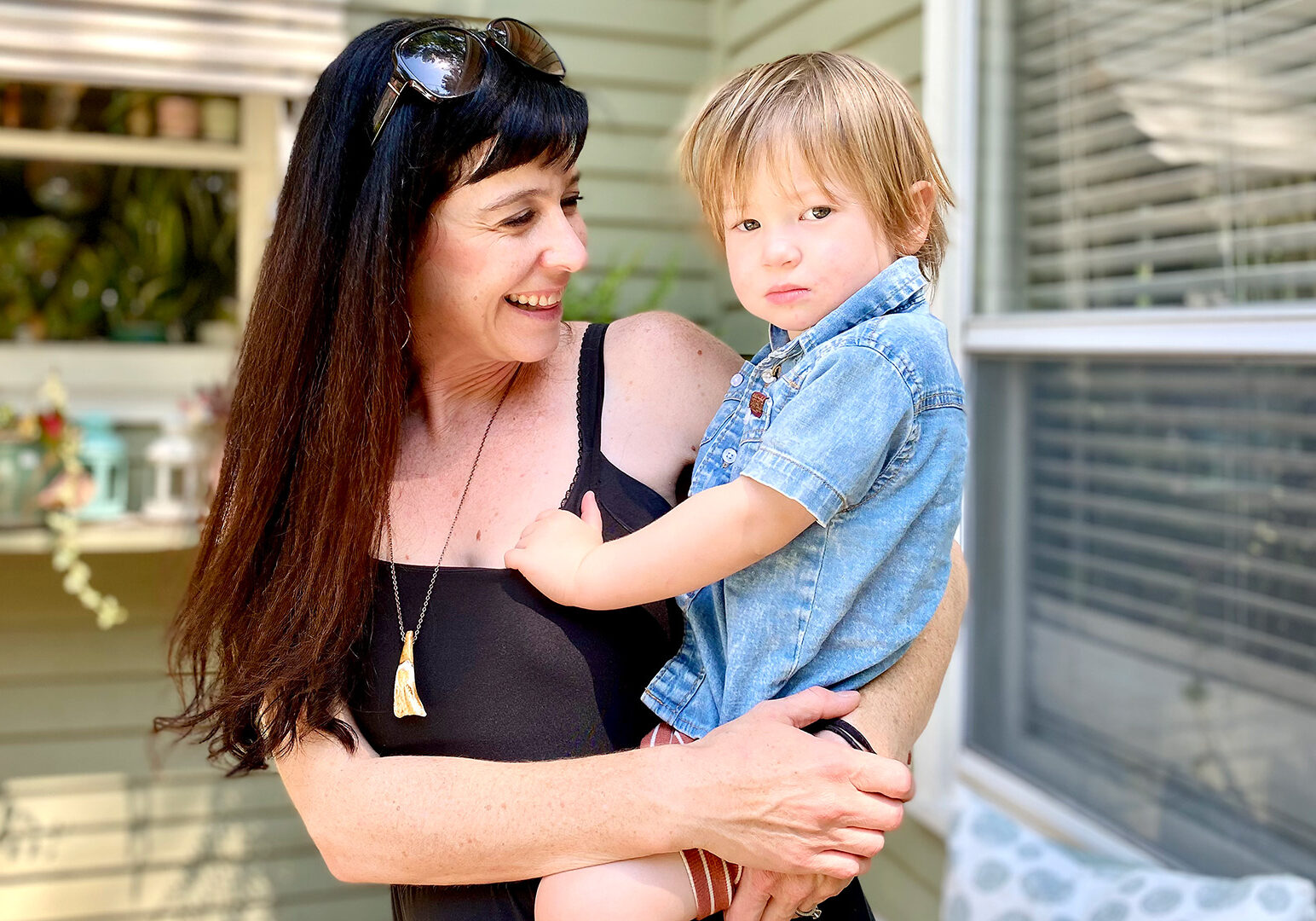 A mother with long brown hair holds her son, who is around two years old, outside of their green home. Lisa Sauciuc was recently able to get her youngest vaccinated from COVID-19.