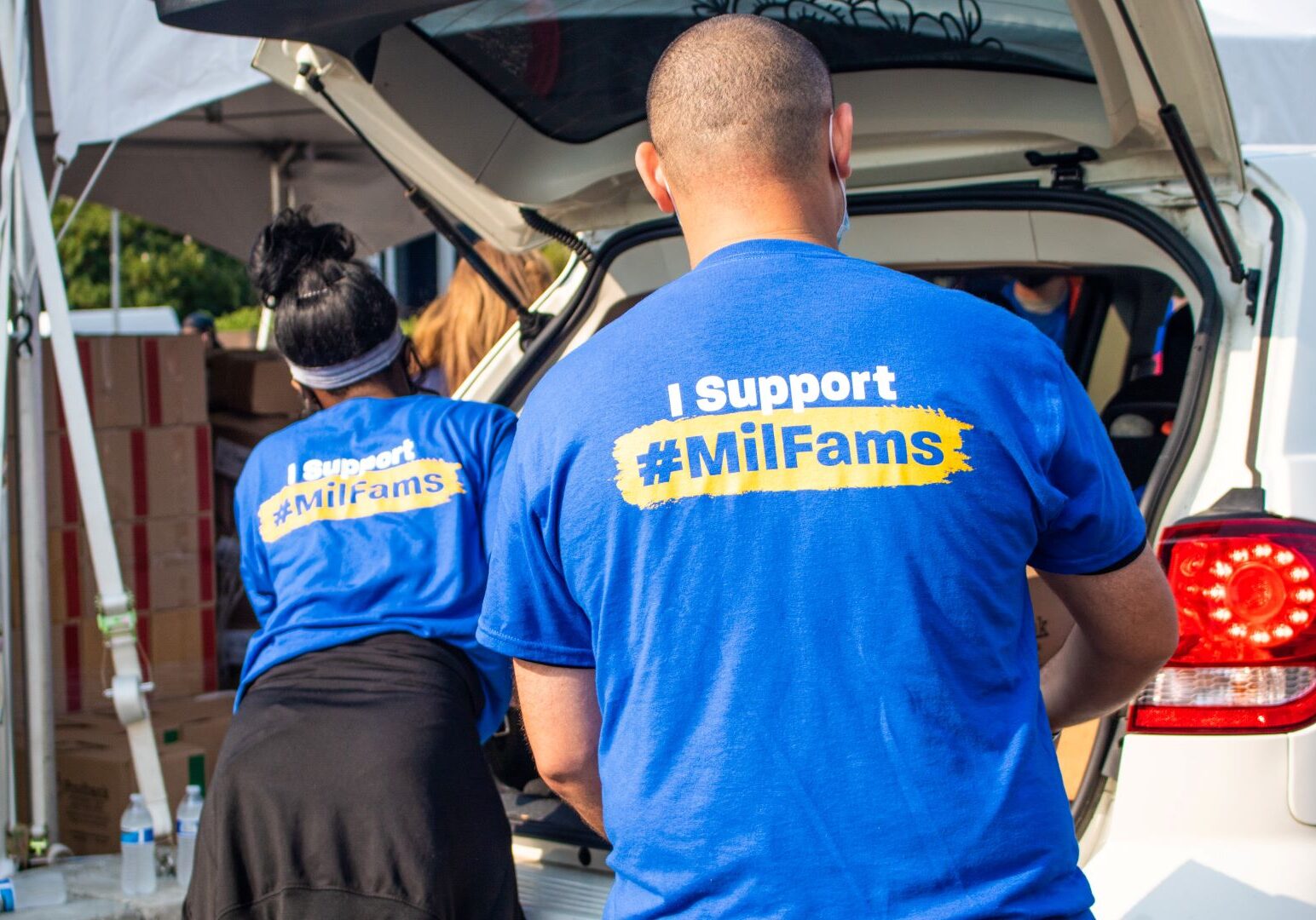 Two people wear shirts that say "I support Mil Fams" and load food into the back of a vehicle.