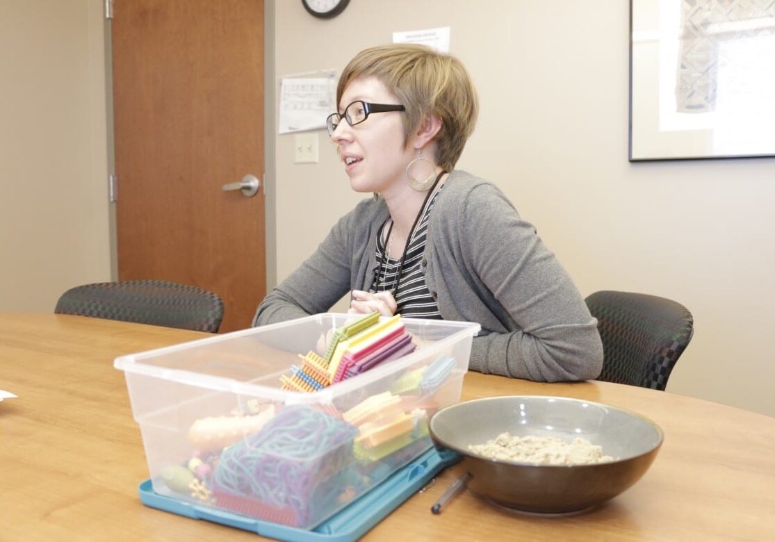 A woman sits at a conference table next to small building blocks and a bowl of sensory sand.