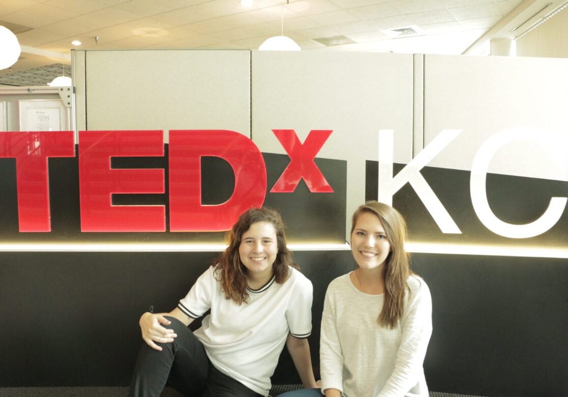 Two girls sit down in front of a TEDxKC sign indoors.