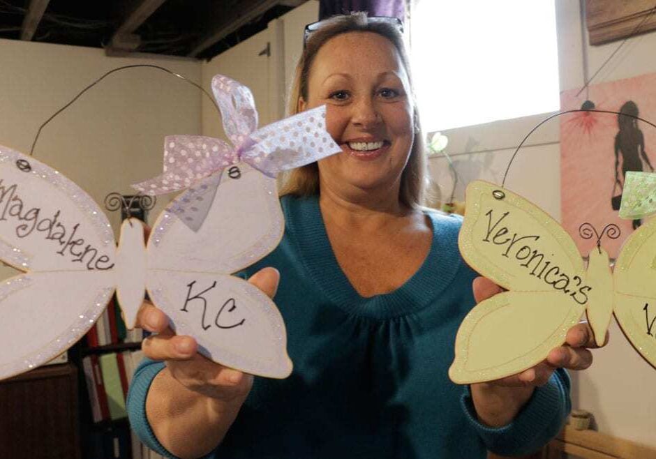 A woman in a blue sweater holds up two decorative butterflies. One says "Magdalene KC." The other says "Veronica's Voice."