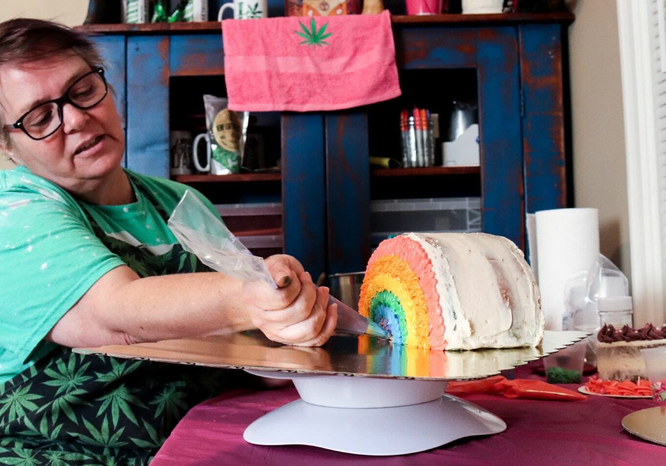a woman with glasses and an apron covered in marijuana leaves holds a piping bag to a cake decorated to look like a rainbow.