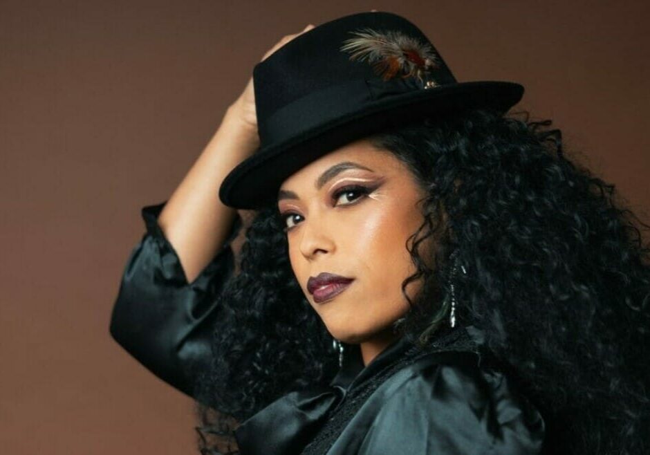 A woman with long curly hair, in a black silk shirt and black fedora hat looks into the camera.