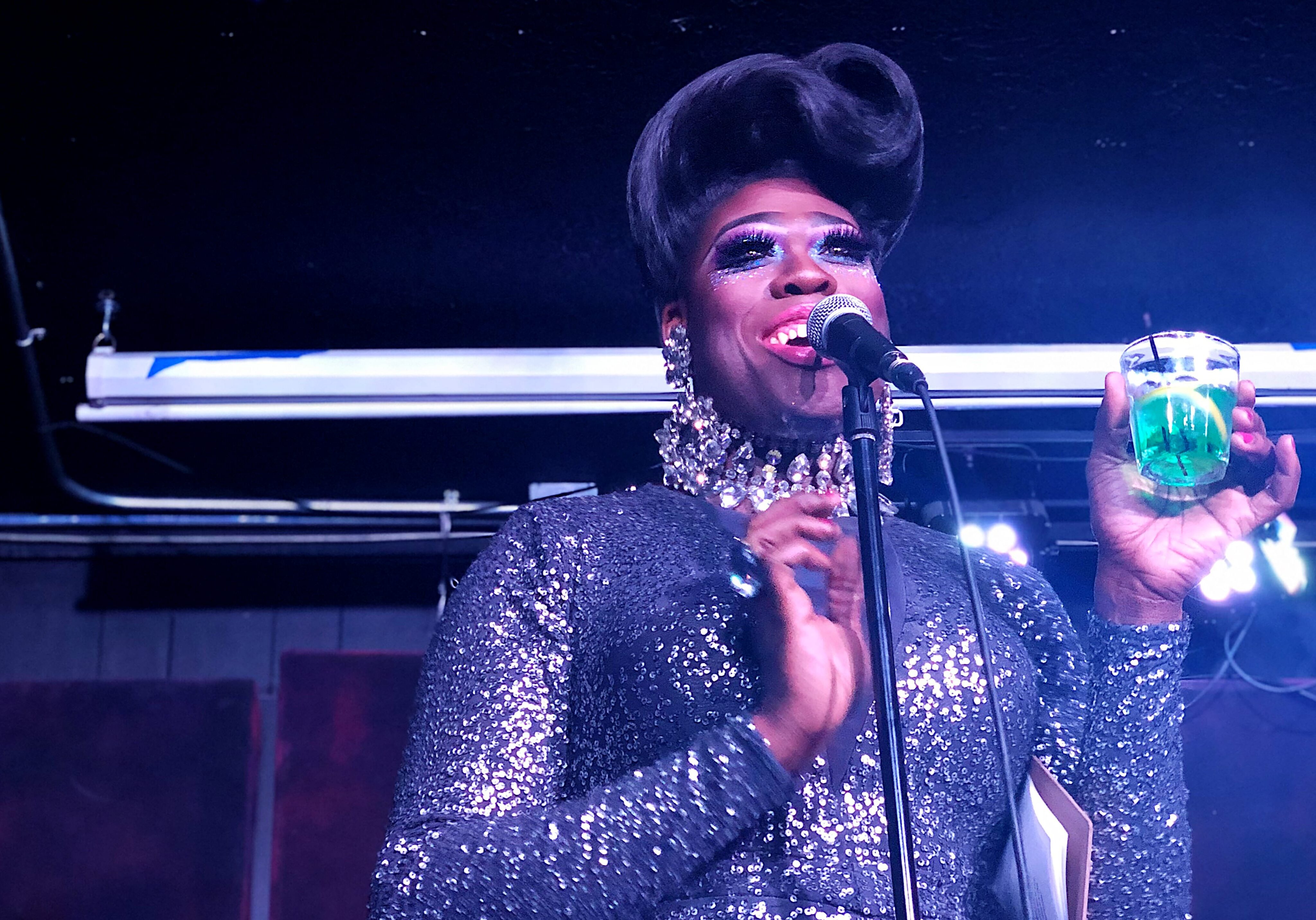 Moltyn Decadence, a drag queen in Kansas City, holds a green drink behind the mic while smiling, in her bouffant and sequin gown