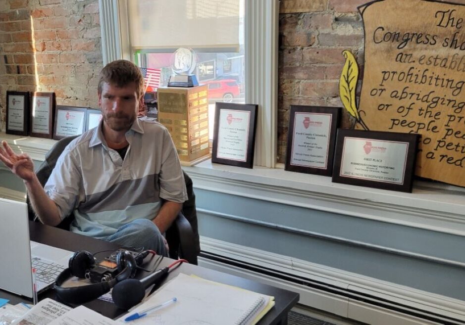 News editor and co-publisher Will Brumleve sits at his desk at the Ford County Chronicle in front of awards and text from the First Amendment. He and co-founder Andrew Rosten launched the newspaper in Paxton, Illinois, in 2020, one of the few new papers in the U.S. in recent years.