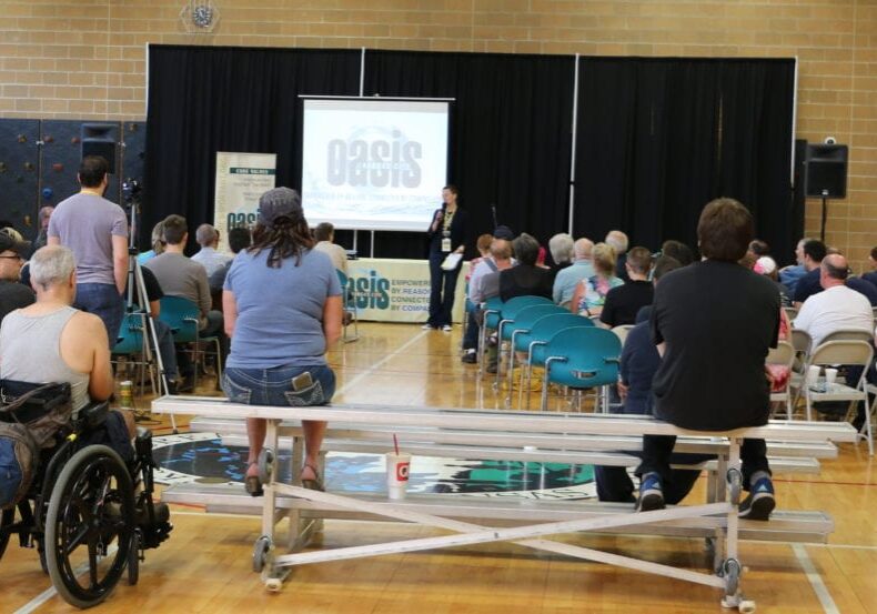 A group of people in a gym sit and listen to a woman speak on the microphone. 
