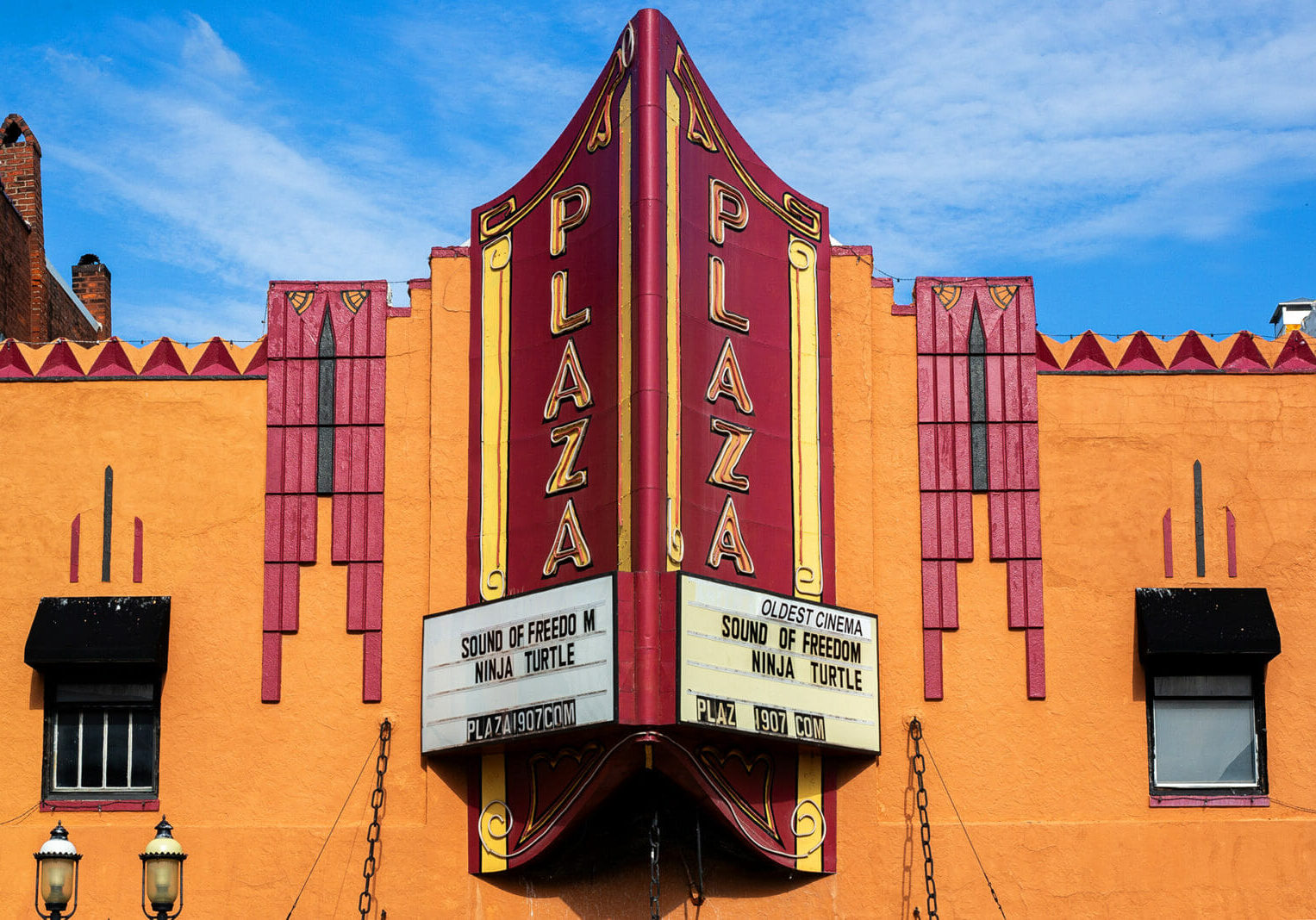 An orange and red building facade shows a big red movie theatre sign that reads "Plaza" on the marquee are movies playing at the theatre and a note reading "oldest cinema"