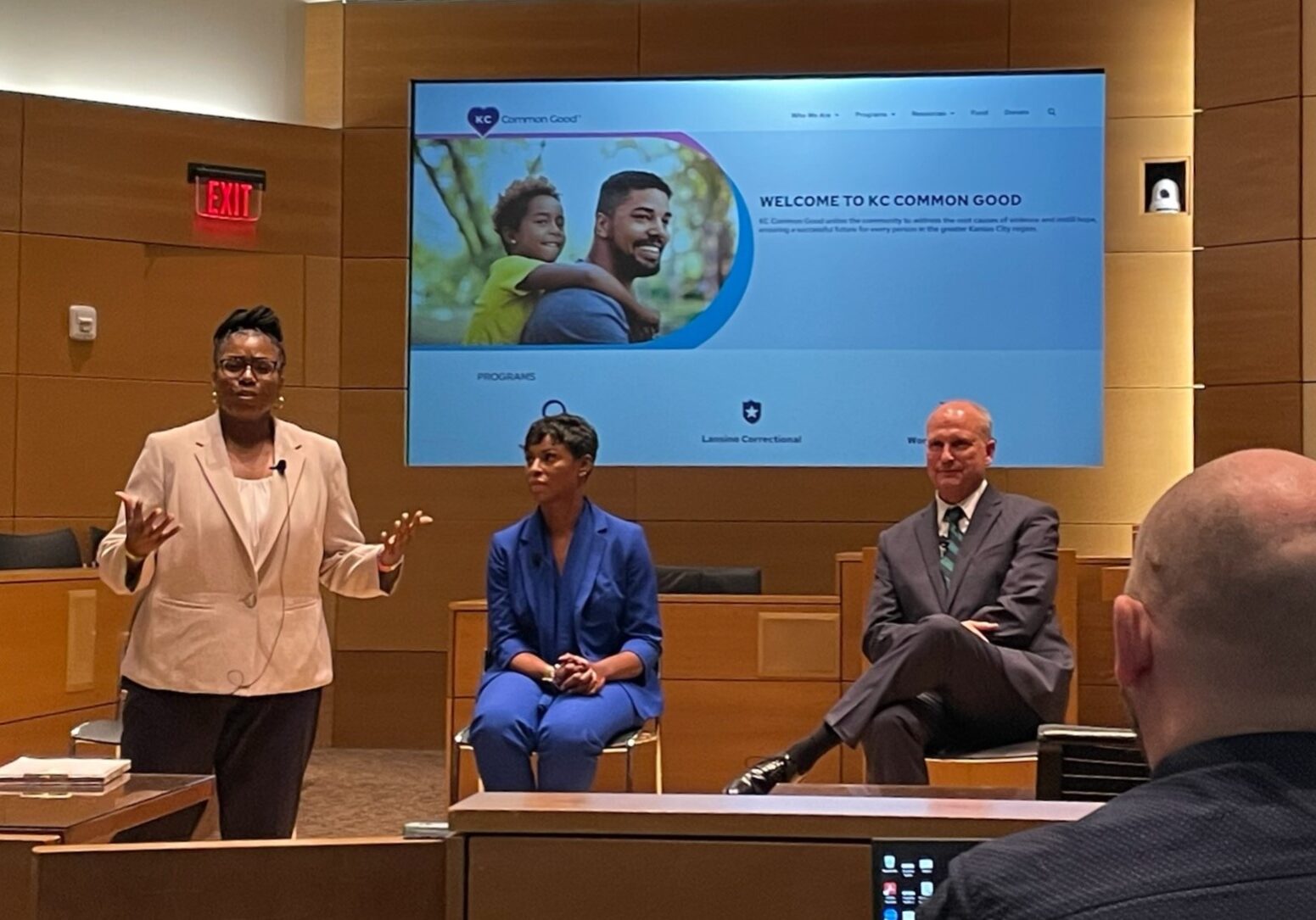 A man, back turned, faces a panel of three people. One woman is standing, and a man and a woman are seated. A large screen behind them.